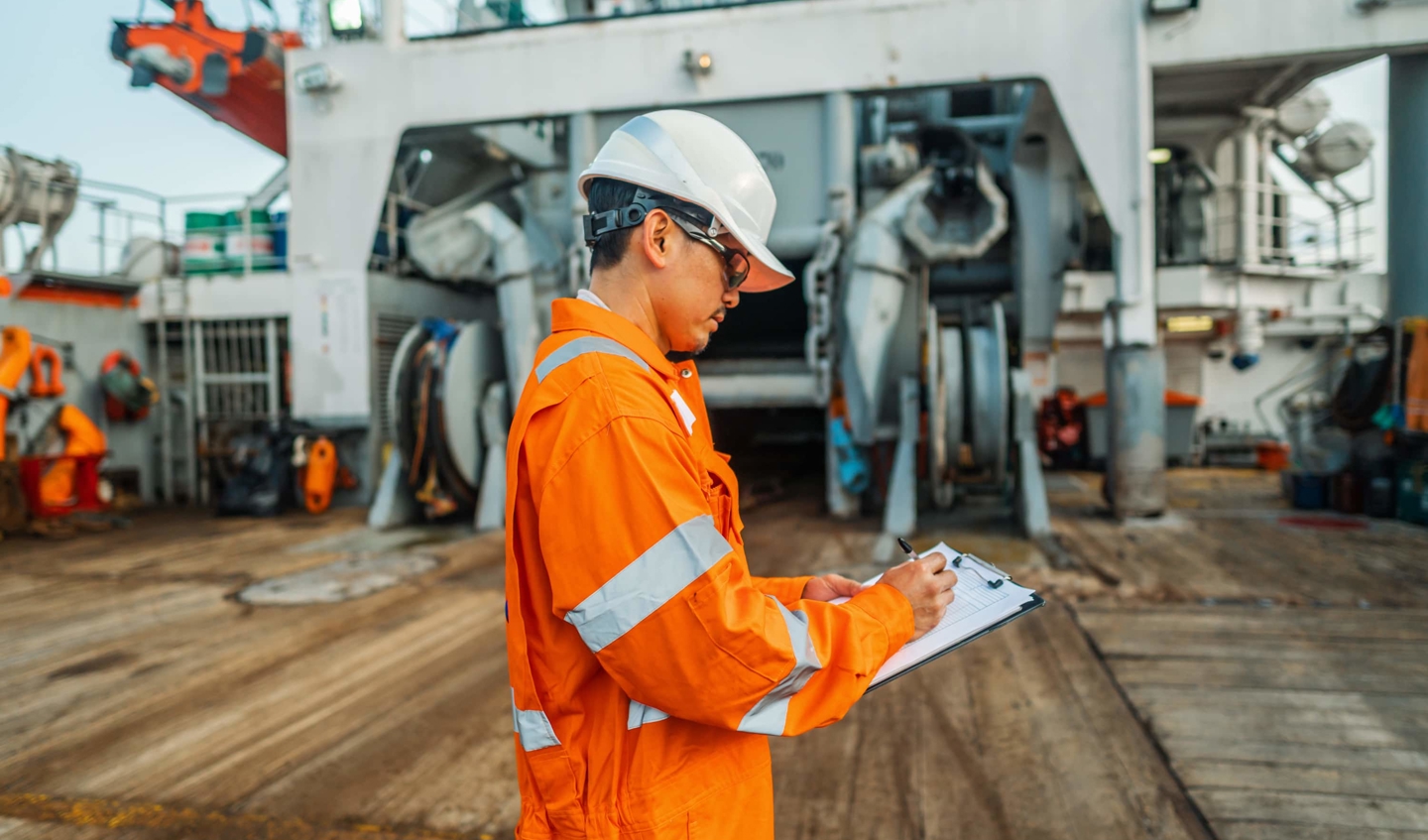 Ship worker in a boilet suit and hardhat looks at a clipboard.