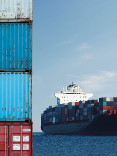 A shipping container sits in the foreground and a ship on the sea in the background.